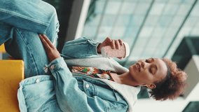 VERTICAL VIDEO, Cute girl with ponytail wearing denim jacket, in crop top with national pattern, drinks morning coffee against the backdrop of modern buildings. Slow motion