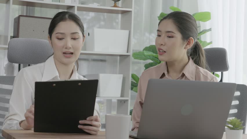 Two young enthusiastic businesswoman working together in the office workspace. Young colleagues having discussion and using laptop together. Modern office workers sitting together at desk.