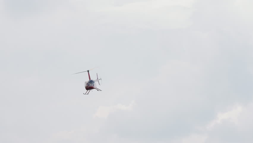 Red american helicopter R44 flying against blue sky background air show. Alone light passenger copter hovering above clouds. Chopper Robinson R-44 fly airshow. Private helicopters travel to America. Royalty-Free Stock Footage #1099005365