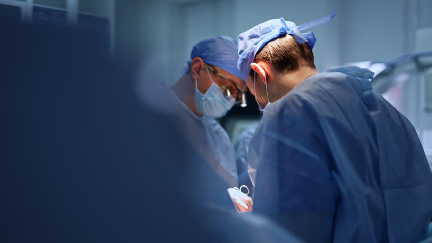 A pair of Caucasian doctors working in surgery room. Focused surgeons operating the patient standing on both sides of him. Blurred backdrop and foreground. Royalty-Free Stock Footage #1099005777
