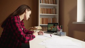 learn virtual. a teenager girl is studying remotely from a teacher online on a laptop sitting at a table next to a bookcase. distance learning education concept. student on exam indoor online