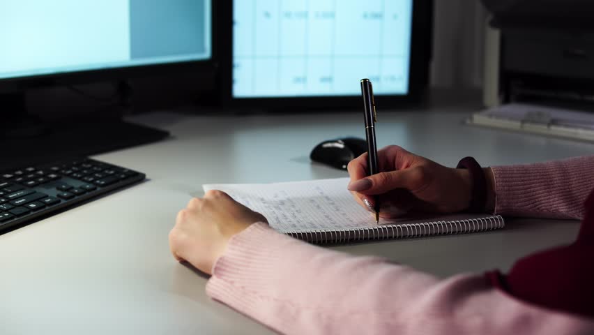 Close up hands of businesswoman holding pen and papers making notes in notepad on table. Financial manager working at office with documents writes information from computer. Employee at workplace.