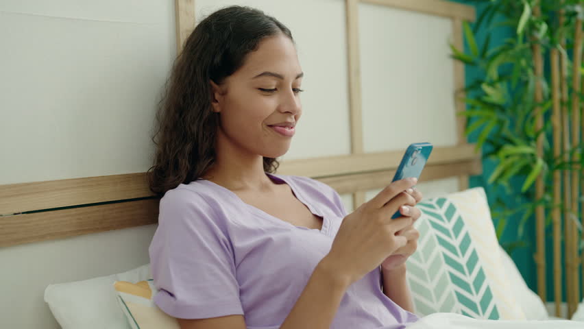 Young african american woman using smartphone sitting on bed at bedroom | Shutterstock HD Video #1099008247