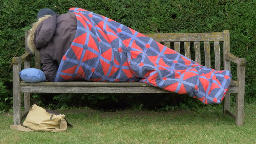 An unidentifiable person wearing winter clothing, having just got into a sleeping bag, lying down during the day on a wooden park bench, with some belongings nearby.