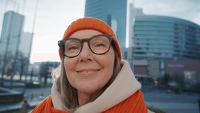 POV positive mature female talking with family via video call on smartphone, record selfie video using mobile phone walking in the middle of urban city landscape. Senior woman, orange winter hat scarf