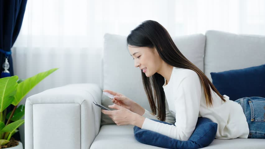 Young asian woman lying on sofa in living room, makes online banking payments through the internet from bank card on smartphone. Shopping online on mobile phone with credit card | Shutterstock HD Video #1099015069