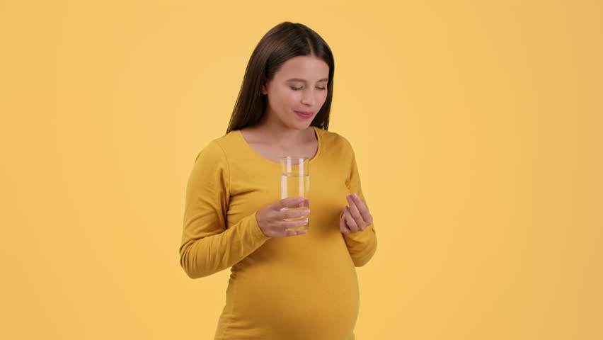 Healthcare during pregnancy. Young pretty pregnant woman taking vitamin pill and drinking water, smiling to camera, orange studio background, slow motion, free space Royalty-Free Stock Footage #1099017957