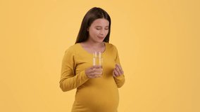 Healthcare during pregnancy. Young pretty pregnant woman taking vitamin pill and drinking water, smiling to camera, orange studio background, slow motion, free space
