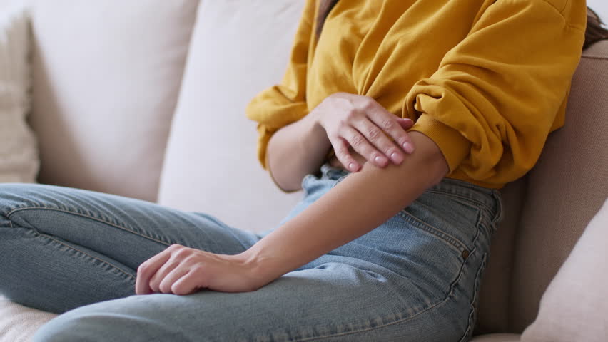 Allergy attack. Close up shot of young unrecognizable woman scratching her hand, suffering from allergic reaction or mosquito bite, sitting on sofa at home, slow motion, empty space