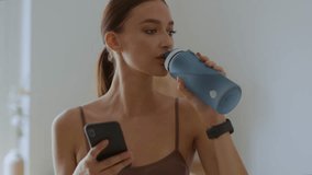 Water balance tracking app. Close up portrait of young sporty woman drinking water from bottle and typing on smartphone, resting after workout at home, tracking shot, slow motion