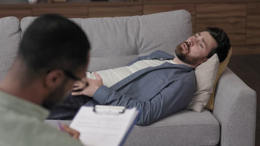 Visit to a psychologist. A handsome man is lying on the couch and talking to a psychotherapist. The patient is in depression, apathy and stress from burnout at work. Help of a psychologist. | Shutterstock HD Video #1099018355