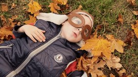 Smiling kid having fun. Boy lying on his back on autumn foliage. Little cute child boy lying on ground and catching falling yellow golden leaves. Child pilot aviator lay on a fallen leaves in forest.