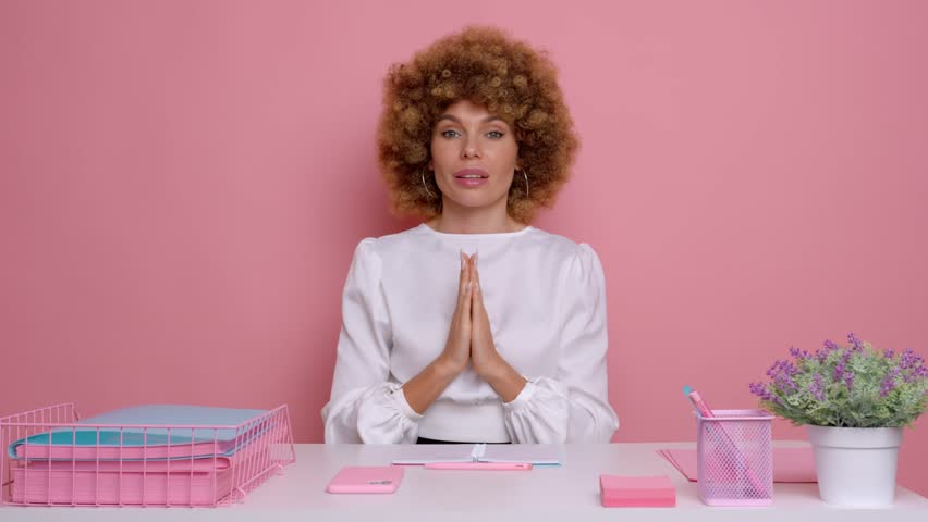 Indoor shot of hopeful young woman employee keeps palms pressed together, sits at work space with papers, has much work to do, isolated over pink background  | Shutterstock HD Video #1099021689