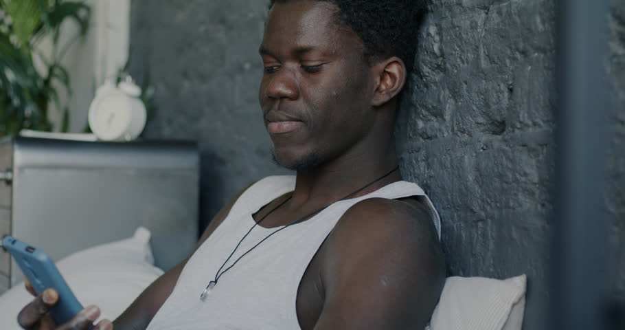 Cheerful African American man speaking on mobile phone relaxing in bed in apartment. Communication and modern technology concept.