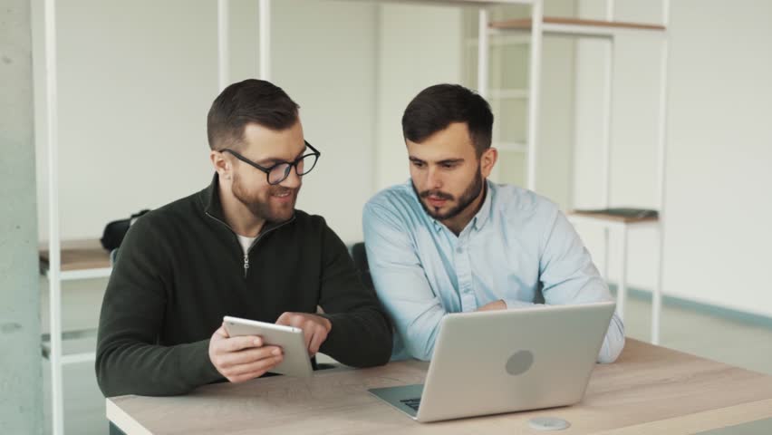 Two male office workers sitting at a laptop and a tablet doing work together | Shutterstock HD Video #1099024701