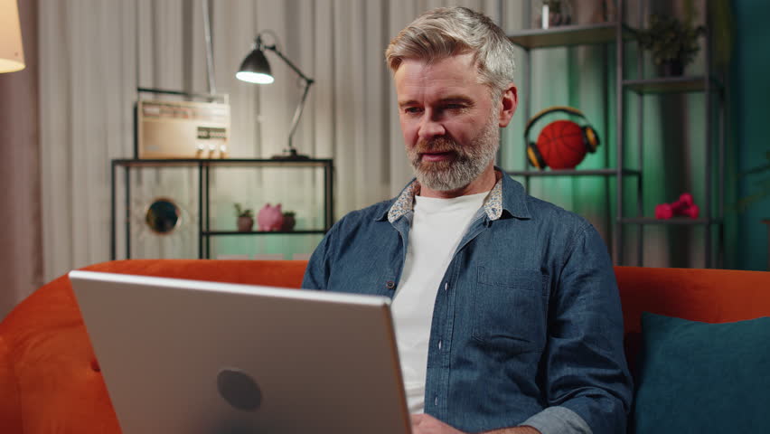 Bearded man sitting on couch closing laptop pc after finishing work in living room at home couch. Mature guy freelancer working online, remote job. E-learning, browsing internet on notebook computer