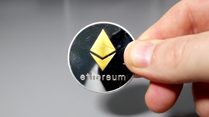Silver and ethereum bitcoin coin held between fingers in hand on gey table | Shutterstock HD Video #1099030785