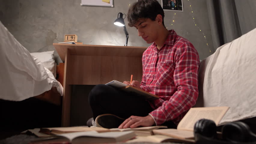 a young Arab student sits at night on the floor in a hostel among open paper books looking for the necessary information. The concept of preparing a homemade creative task. Royalty-Free Stock Footage #1099031143