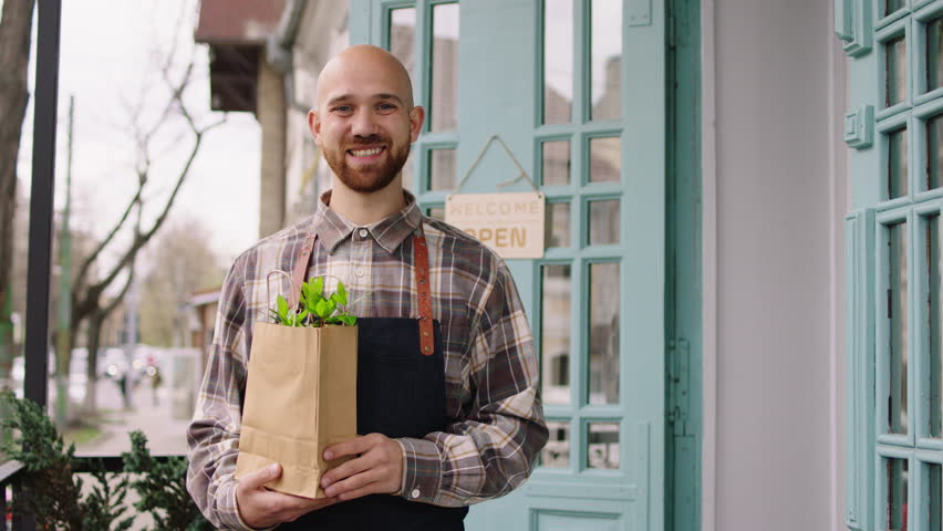 Cute floral shop owner and florist man smiling large in front of the camera while holding a eco bag with some plants inside beside of the shop