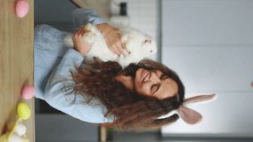 Vertical video. Portrait of the smilling woman holding the fluffy easter rabbit, wearing bunny ears. Smiling lady is getting ready for the spring holiday. Preparation for easter