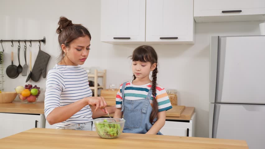 Caucasian mother teach and motivate young daughter eat green vegetable. Adorable child shake head say no to mom, don't want to eat salad on dinner plate in kitchen. Healthy food and childhood concept. Royalty-Free Stock Footage #1099040845