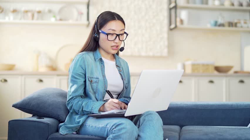 Young Asian woman in a headset and with a laptop communicates online, writes in a notebook while sitting on the sofa in the living room. Female student talking via video call at home on couch indoor | Shutterstock HD Video #1099042815