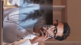 Vertical video. Patient diagnosis. Male doctor. Mobile consultation. Expressive man talking smartphone holding x-ray photo of lungs sitting work desk light room interior.