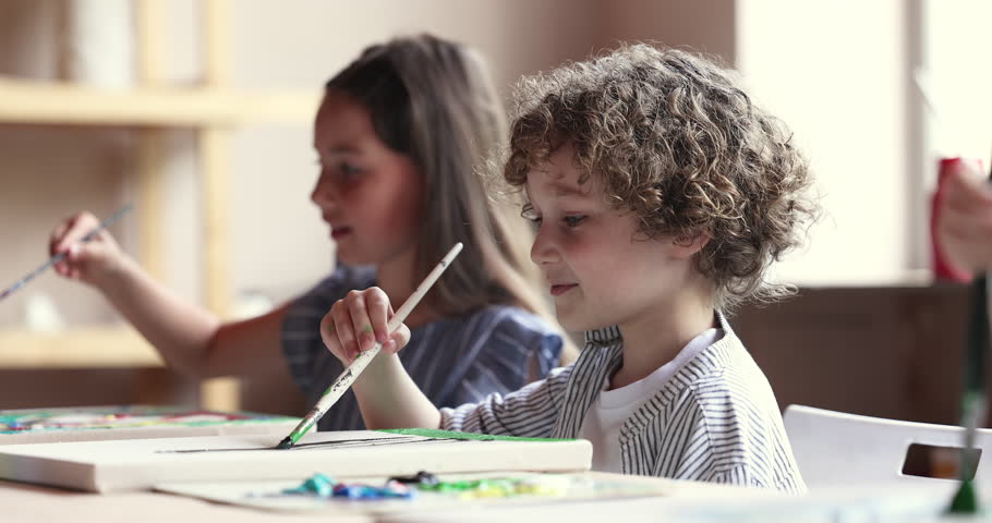 Pretty curly-haired little boy holding paintbrush painting on canvas seated at table with other preschoolers, art class participants. Children talents development, creative hobby, day-care activity Royalty-Free Stock Footage #1099050105