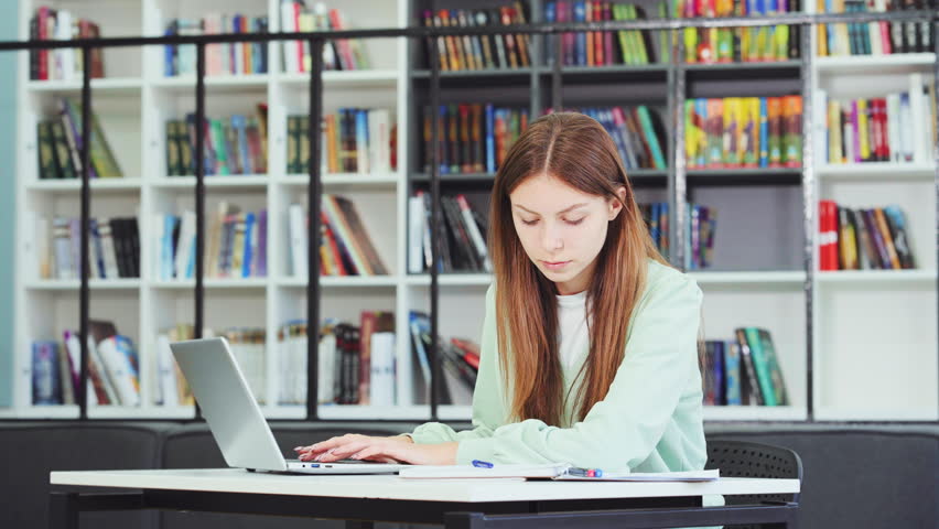 Young attractive university student studying at library. Caucasian teenager writing notes using laptop, planning working process. Exam preparation