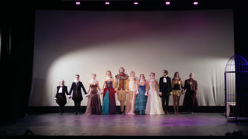 Group of performers holding hands walking on stage bowing and clapping. Wide shot portrait of talented Caucasian actors and actresses in elegant retro-style costumes thanking in theater after show