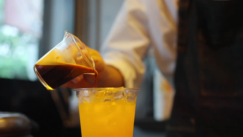 Asian man barista making iced black coffee with orange juice in takeaway glass. Male coffee shop waiter serving coffee to customer on counter at cafe. Small business cafe and restaurant owner concept.