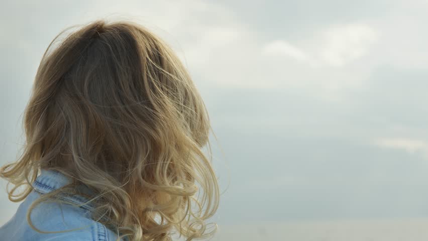 close up portrait of blonde girl with curly hair turns her head against backdrop of sky. child in denim jacket with hair blowing in wind looks to side.