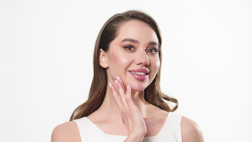A slender young beautiful caucasian woman with dark hair in a white top turns to the left, touching her jaw line and smiling broadly at the camera on a white background. Moisturizer ad | Shutterstock HD Video #1099065357