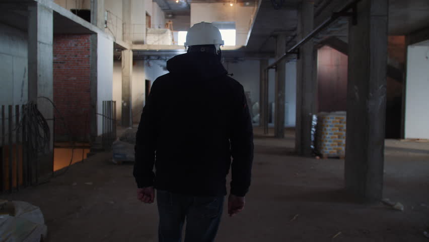 Worker in helmet walks inside unfinished building at construction site backside view. Inspector in future house