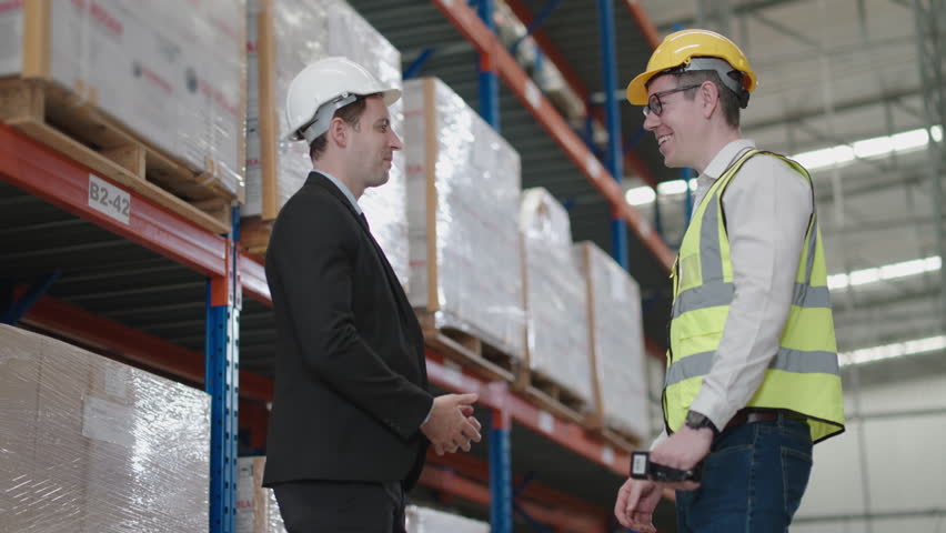 businessman shaking hands with worker wearing hardhat standing against warehouse shelves in background.worker wearing reflective jacket holding QR code scanner standing in factory warehouse smiling.