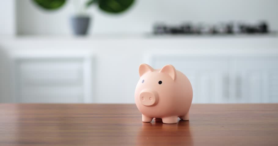 on the wooden table in kitchen is a piggy bank in a bright space. A woman's hand puts a coin in an peachy open piggy bank. The concept of saving money in a piggy bank or new start up. straight view. Royalty-Free Stock Footage #1099077211