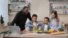 Young attractive multiracial family preparing meal from fresh vegetables. Cute preschool children feeding with salad african dad and charming mom cooking dinner together in their in kitchen.
