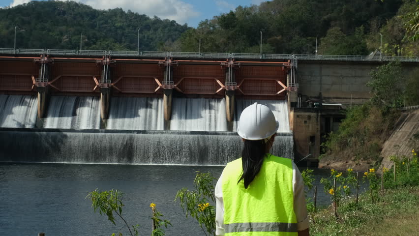 Female engineer wearing green vest and white helmet working outside dam with hydroelectric power plant and irrigation. Renewable energy system. Sustainable energy concept.