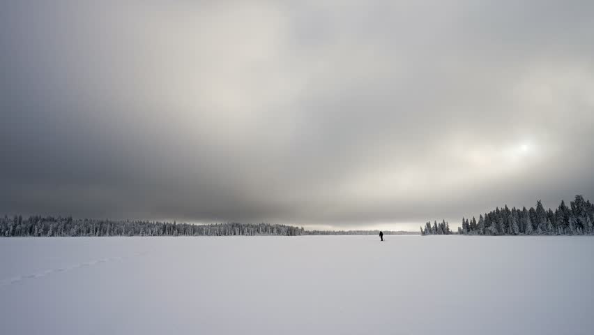Person in the distance wearing black id snowshoeing across a frozen lake with a shoreline of spruce trees under a gray sky. The sun can be seen through the thin clouds.
