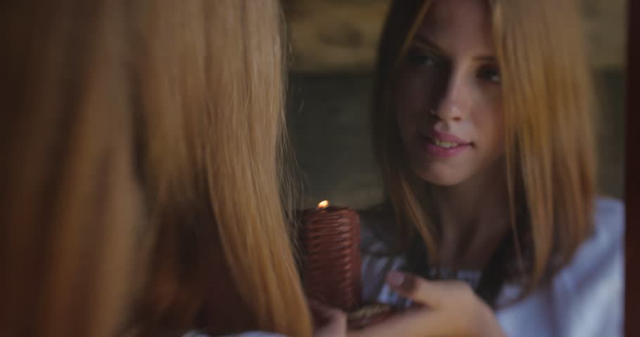 Close up attractive young blonde woman in an embroidered shirt looking into a mirror in an old wooden house, smiling and holding a candle. Divination, mysticism, love magic concept. | Shutterstock HD Video #1099090547