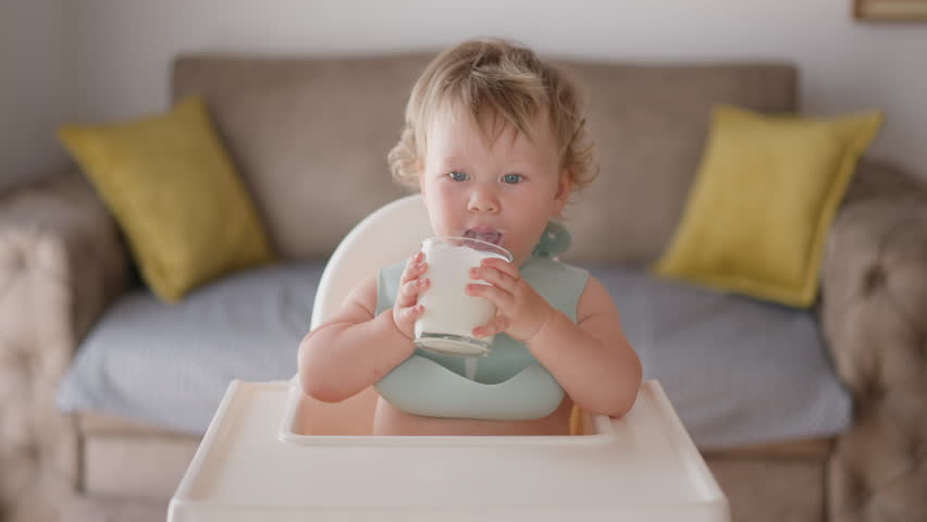 Little mixed race boy child drinking milk yogurt. Healthy eating toddler having breakfast in living room. Daughter girl holding glass with kefir in baby chair and licks her lips. Positive kid smiling. | Shutterstock HD Video #1099102697