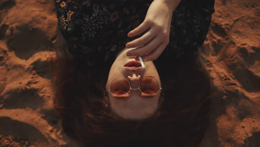 Portrait of young hippie woman with closed eyes smoking lying on sand beach at sunset. Female model wearing sunglasses smoke looks at camera, dramatic hipster girl. Teenage lady with red curly hair