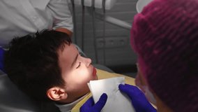 Close-up of a teenage boy on dentist's chair getting teeth treatment in modern pediatric dentistry clinic. Rear view of a dentist hygienist wiping mouth of a little patient after dental procedures.