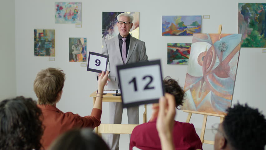 Middle age auctioneer with gray hair calling out bids while selling contemporary painting on art auction Royalty-Free Stock Footage #1099108333