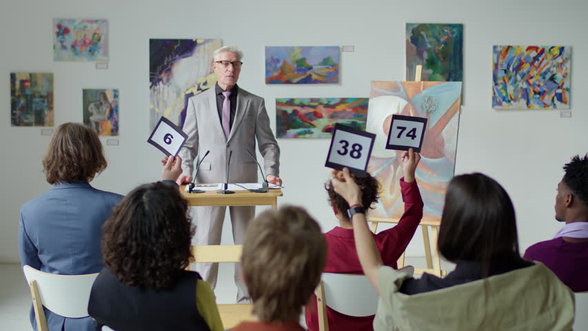 Male auctioneer with gray hair standing by rostrum on stage and calling out bids while selling artwork on auction Royalty-Free Stock Footage #1099108615