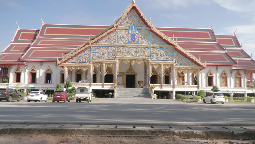 A stationary footage of a travelling cyclist couple that passes by a large sacred Asian temple at the city of Udon Thani in Isan, Thailand. The guy stares at it as he drives by.