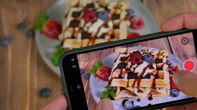 Food blogger recording a video of sweet dessert, woman using smartphone, taking a photo of pile of soft fluffy waffles served with fresh berries and chocolate sauce, food photography, 4k video footage
