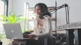 Young optimistic attractive African American woman teacher records video lesson for students and school children want to gain additional knowledge via Internet sits on sofa near laptop and microphone