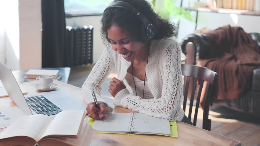 Young happy African American woman in headphones listens to internet podcast and laughs making notes in workbook gets quality education and opportunity to change careers sits at table in home interior | Shutterstock HD Video #1099116445