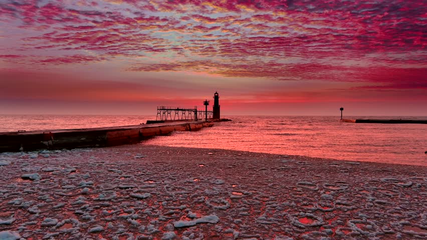 Iconic Lighthouse beacon silhouetted against fiery Lake Michigan twilight horizon, moving aerial view.